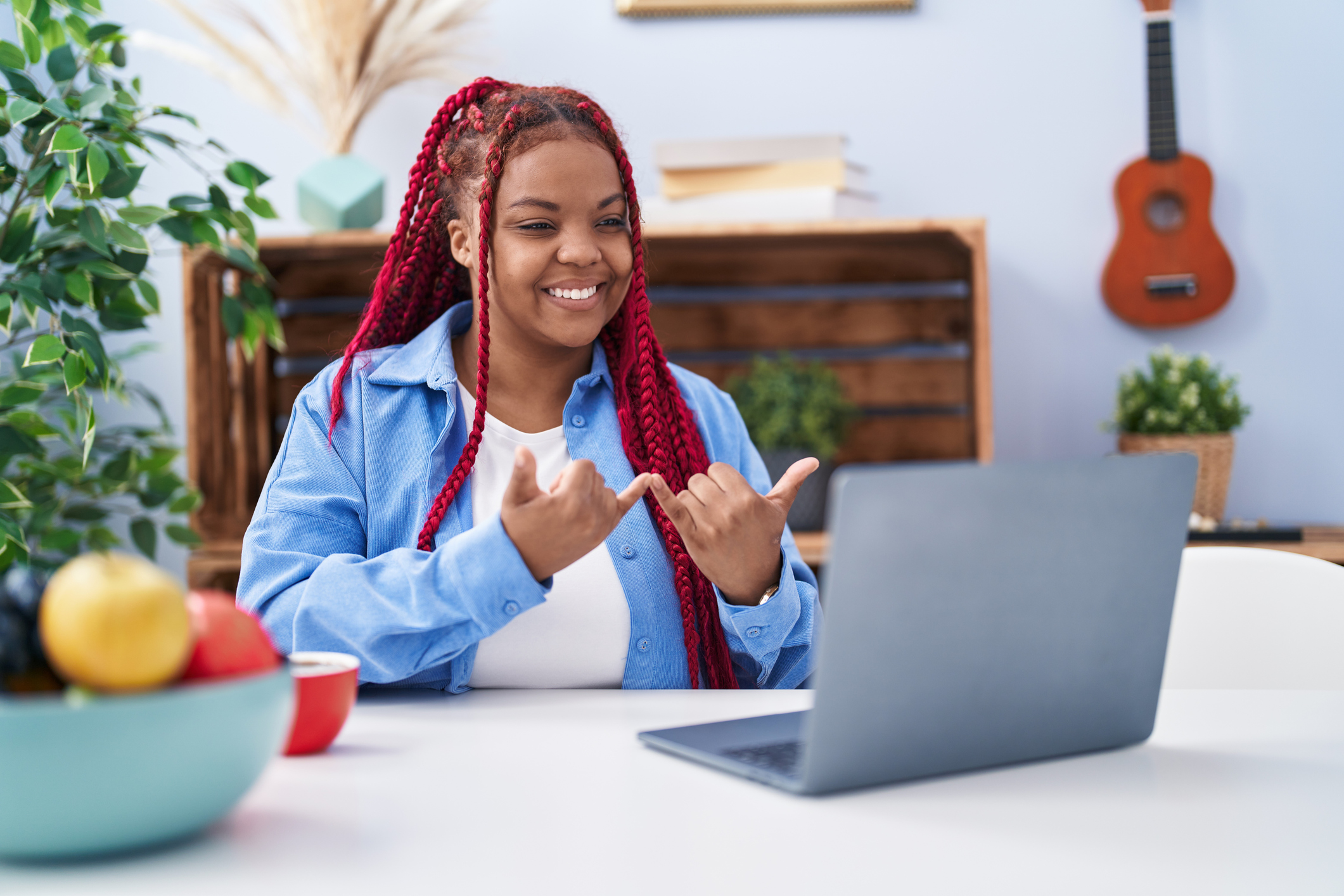 A Black woman with red braids using sign language online while sitting at a kitchen table