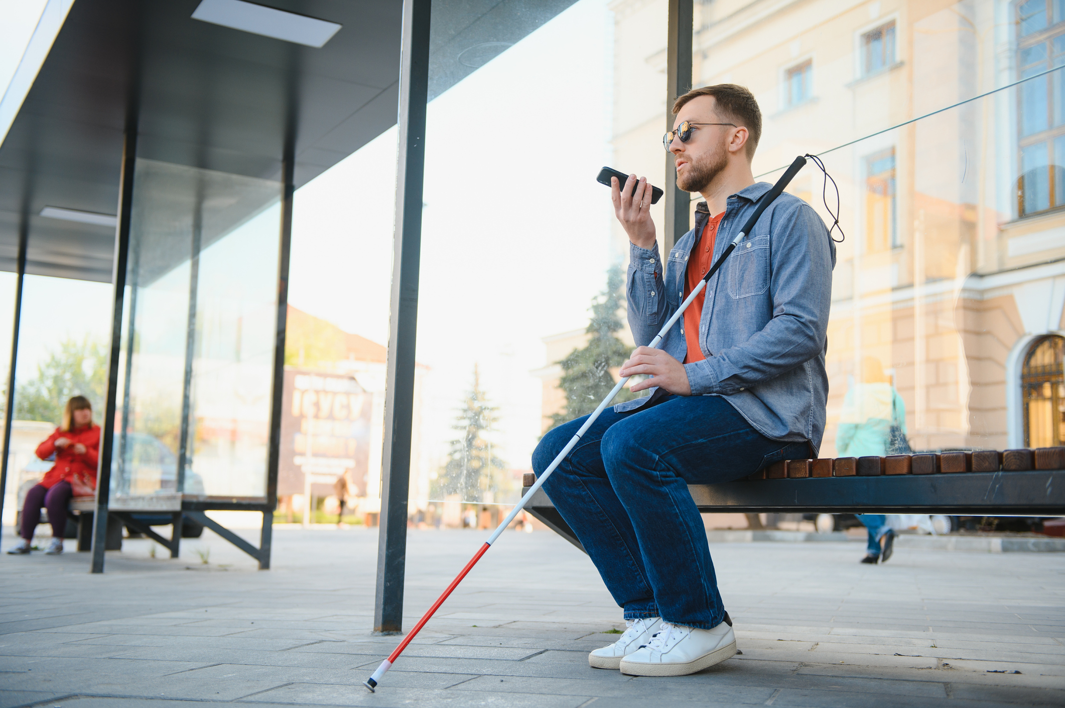 A man who is blind or low vision sits on a bench and uses a mobile device