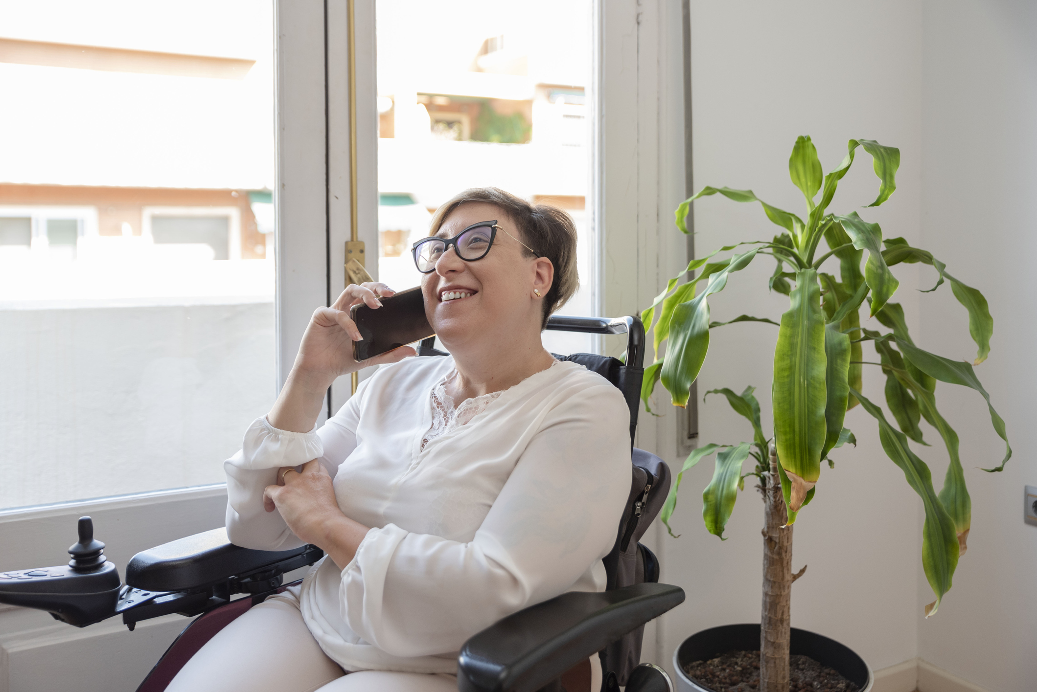 Happy businesswoman in wheelchair sitting next to window talking on smartphone