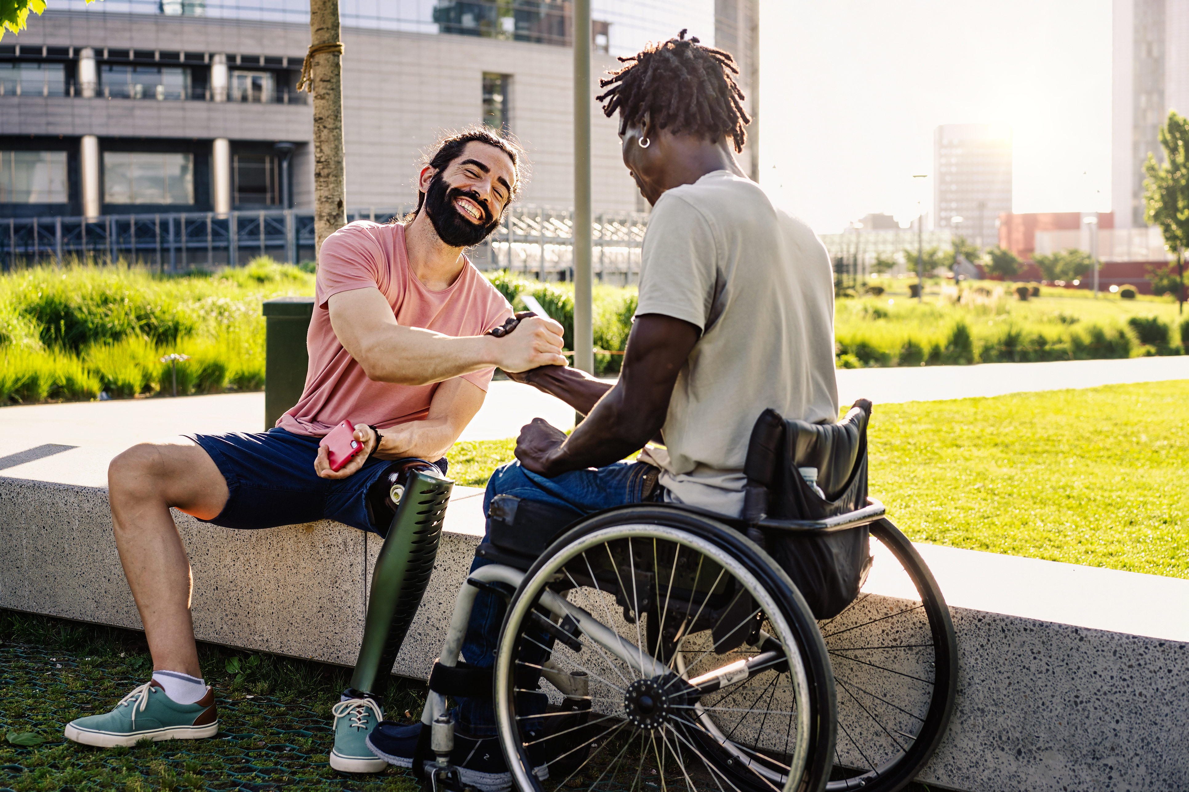 A Latino man with an artificial leg prosthesis shakes hands with a Black man sitting in a wheelchair