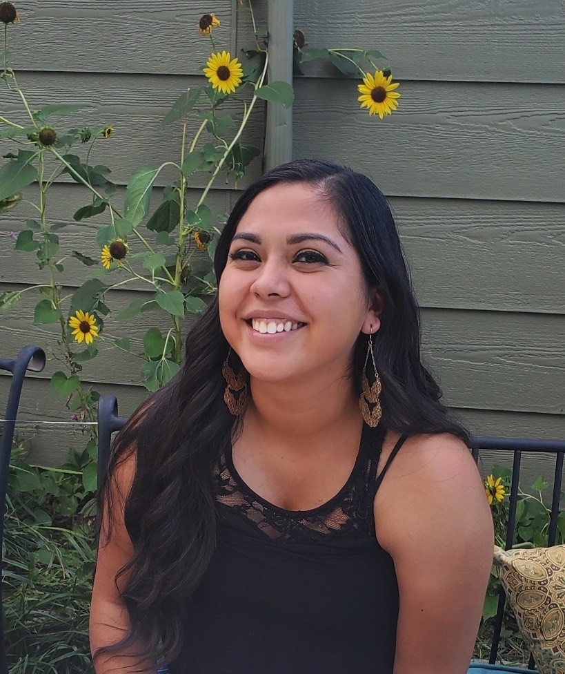Sheena Leal with long, dark hair and a bright smile sits on a bench outdoors. She is wearing a black tank top with a lace detail and long, brown, patterned earrings. Behind her is a light green wall and several tall sunflower plants with yellow blooms.