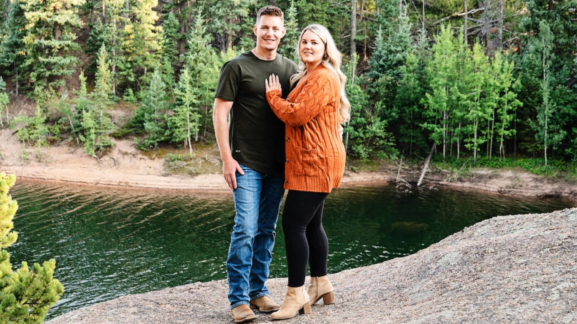 Couple standing together in front of a lake with trees in the background.