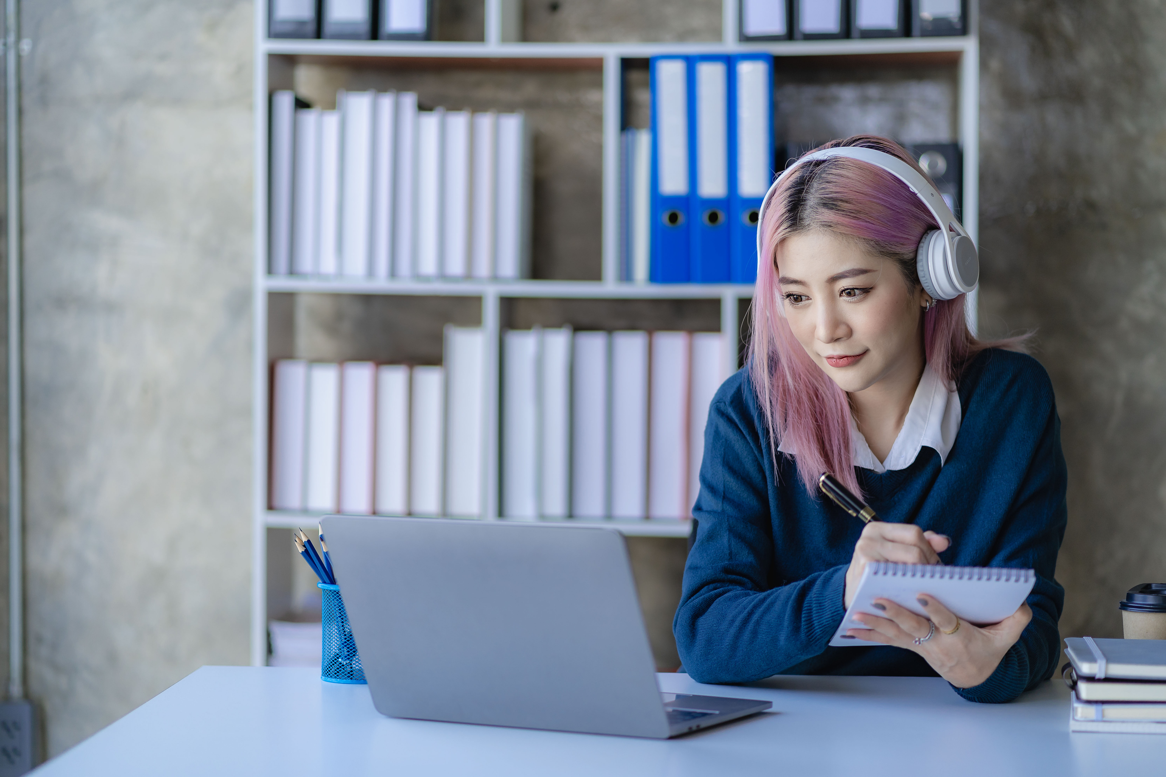 Asian female college student with pink hair wears headphones and uses a smartphone and laptop