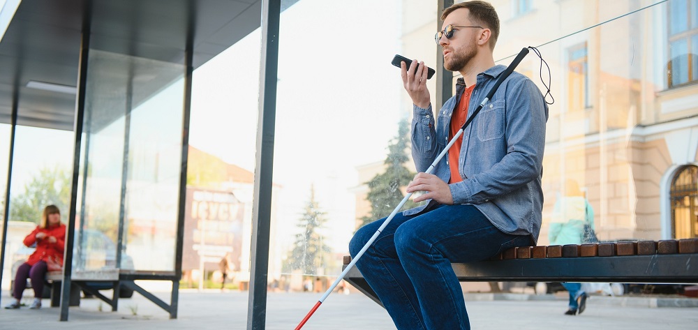 A man who is blind or low vision sits on a bench and uses a mobile device