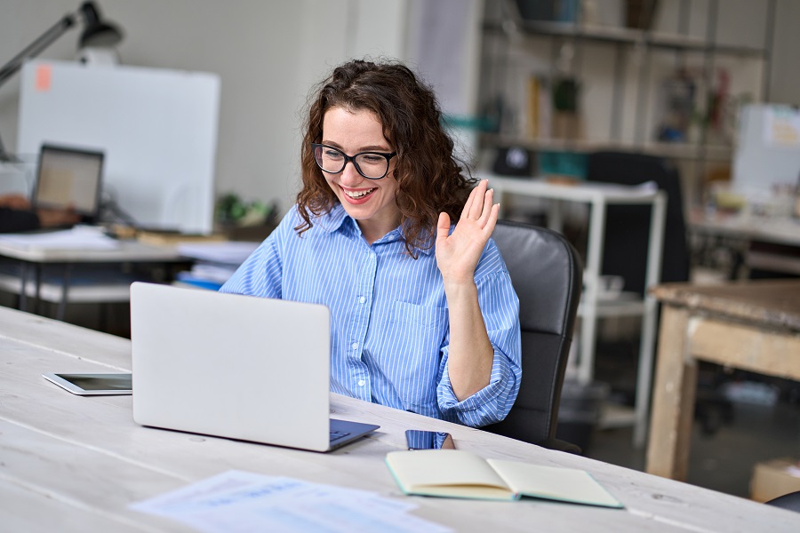 Smiling business woman in an office waving at a laptop screen and having a virtual work meeting
