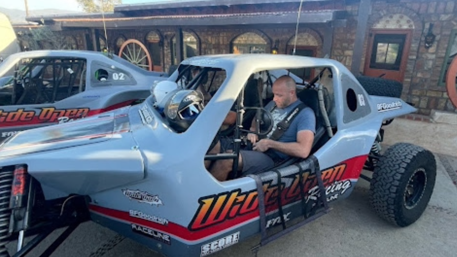 A man sits in a grey off-road racing car, buckling his harness with a helmet, waiting, with another racing car parked beside him in front of a stone building under a clear blue sky.