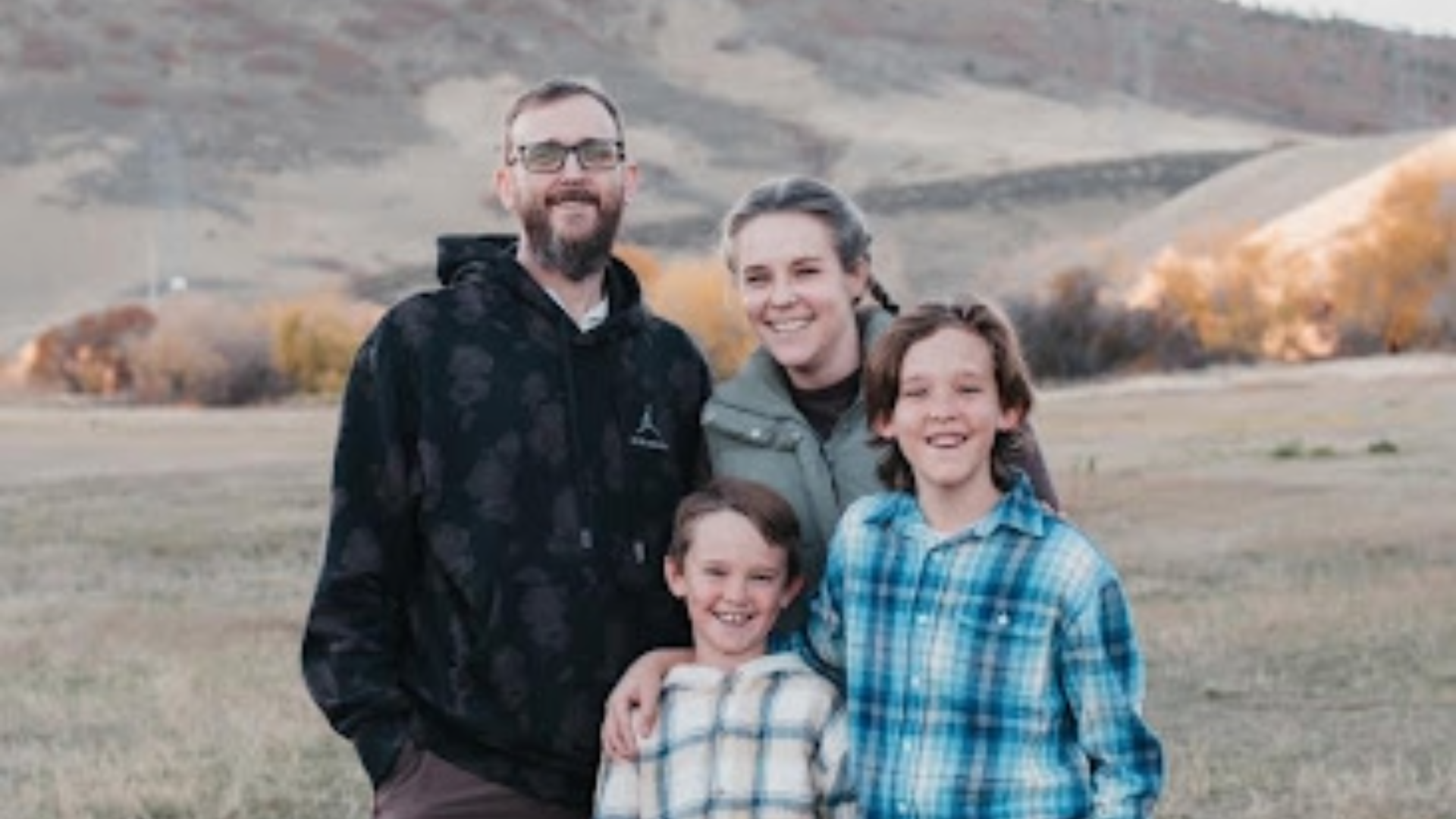 A high-angle outdoor portrait of a family standing in a dry, autumnal field. The background features rolling hills under a soft, overcast sky. Everyone is dressed in casual, cool-weather clothing