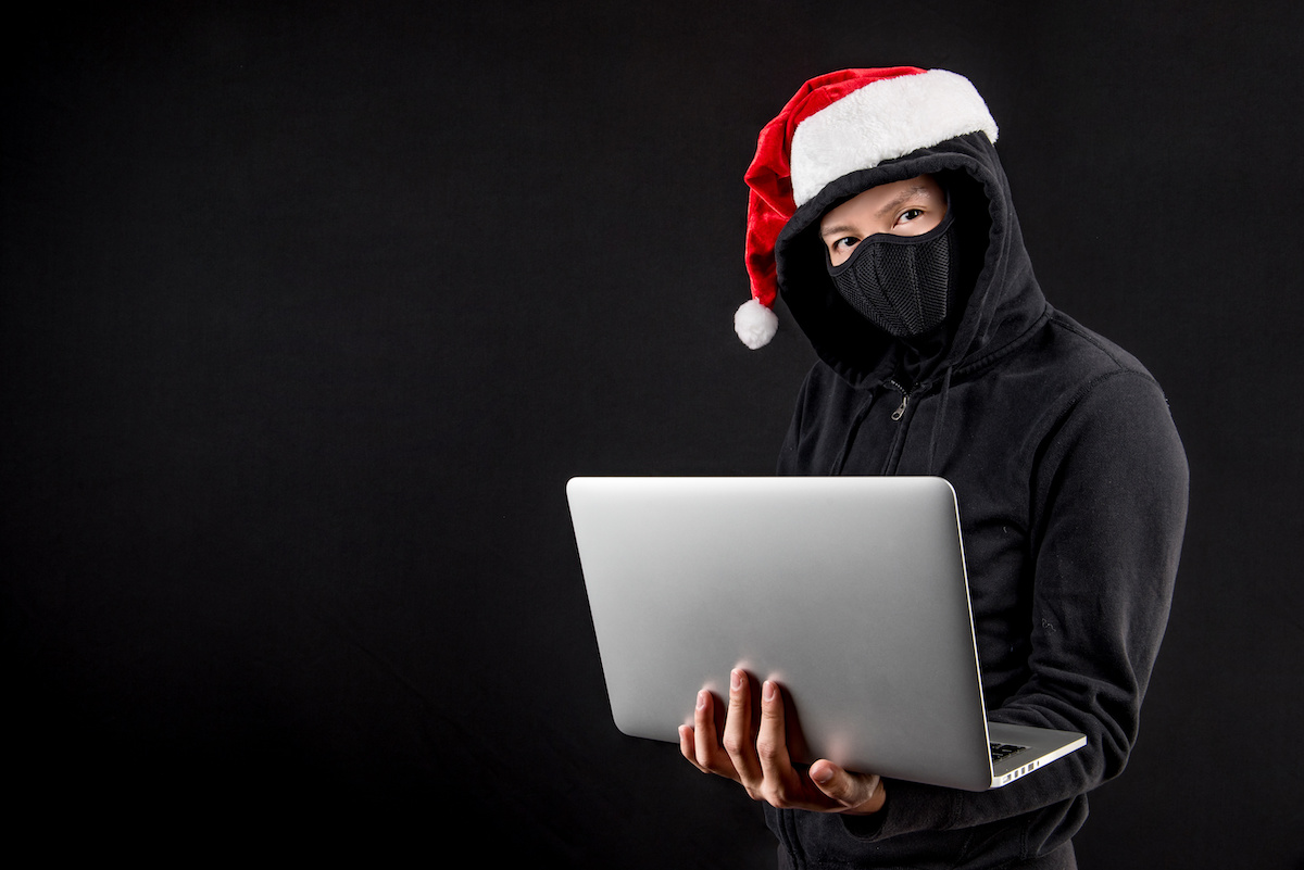 Person wearing all black clothing, a black mask and a Santa hat while holding a laptop