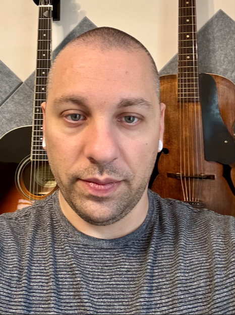 A medium close-up selfie of Lane Sidebottom with a short, gray and black striped shirt looking directly at the camera with a neutral expression. He is wearing small white earbuds and stands in front of a gray sound-dampening wall. Two acoustic guitars are visible, one on the left and one on the right, partially cut off by the frame.