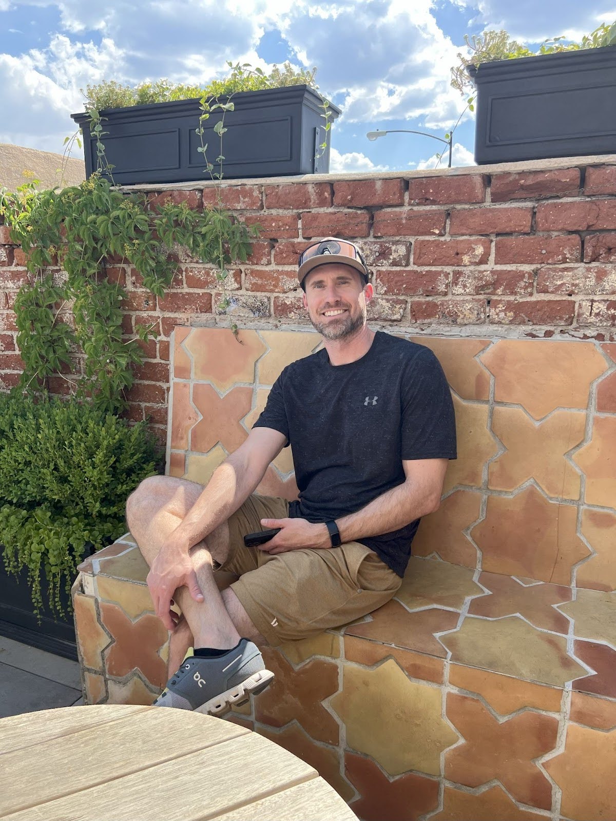 Sean Clark with a beard and a baseball cap is seated on a tiled bench outdoors, smiling at the camera. He's wearing a dark t-shirt, khaki shorts, and athletic shoes. Behind him is a brick wall with planters containing greenery, and to his left, there's a lush green bush. A wooden table is partially visible in the foreground. The sky is bright with some clouds.