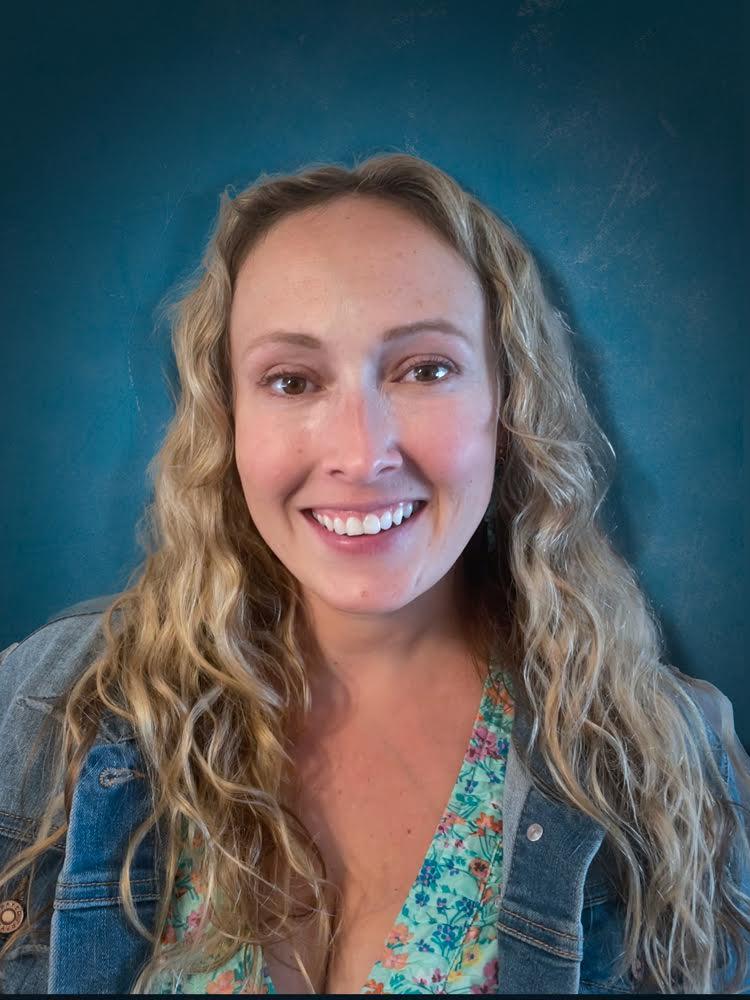 A medium skinned toned woman with blond slightly curly long hair, brown eyes, smiling, wearing a jean jacket and a floral undershirt.