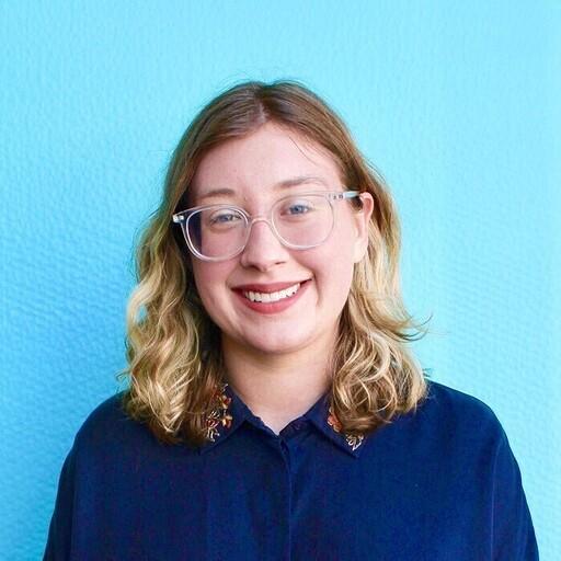 A profile photo of Abigail Fisher. Abigail is a person who is white with shoulder-length blonde, curly hair and clear-framed glasses. She has a dark blue, collared long-sleeve shirt on and is standing in front of a light blue wall.