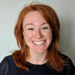 A profile image of Alex Orlov. Alex has red, medium-length hair and a dark blue shirt. She is standing in front of a grey wall.