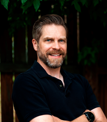 Man with dark brown hair and beard standing with his arms crossed in a black t-shirt smiling. 