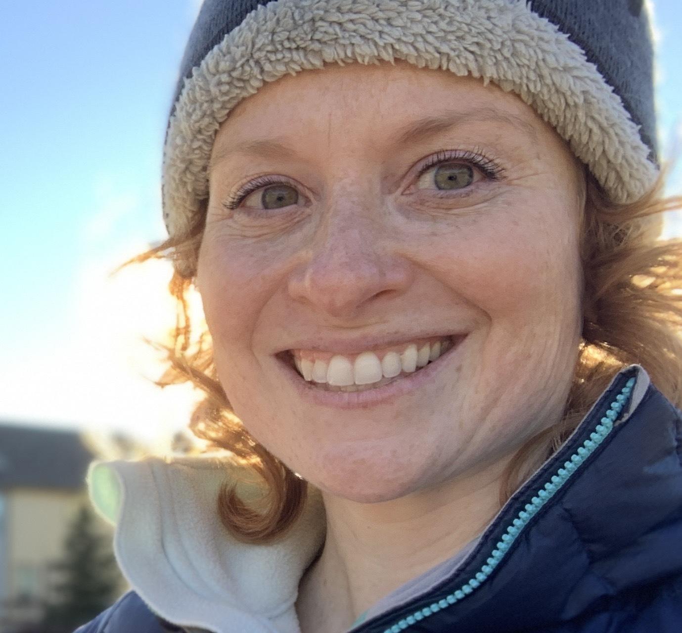 A profile photo of Debra Alban. Debra is a person who is white with shoulder-length, curly red hair. She is wearing a fur hat and winter jacket and is standing in front of a blue sky.