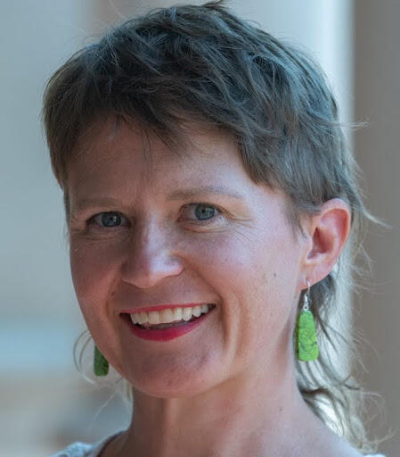 Woman with short light brown hair wearing bright green earrings smiling.