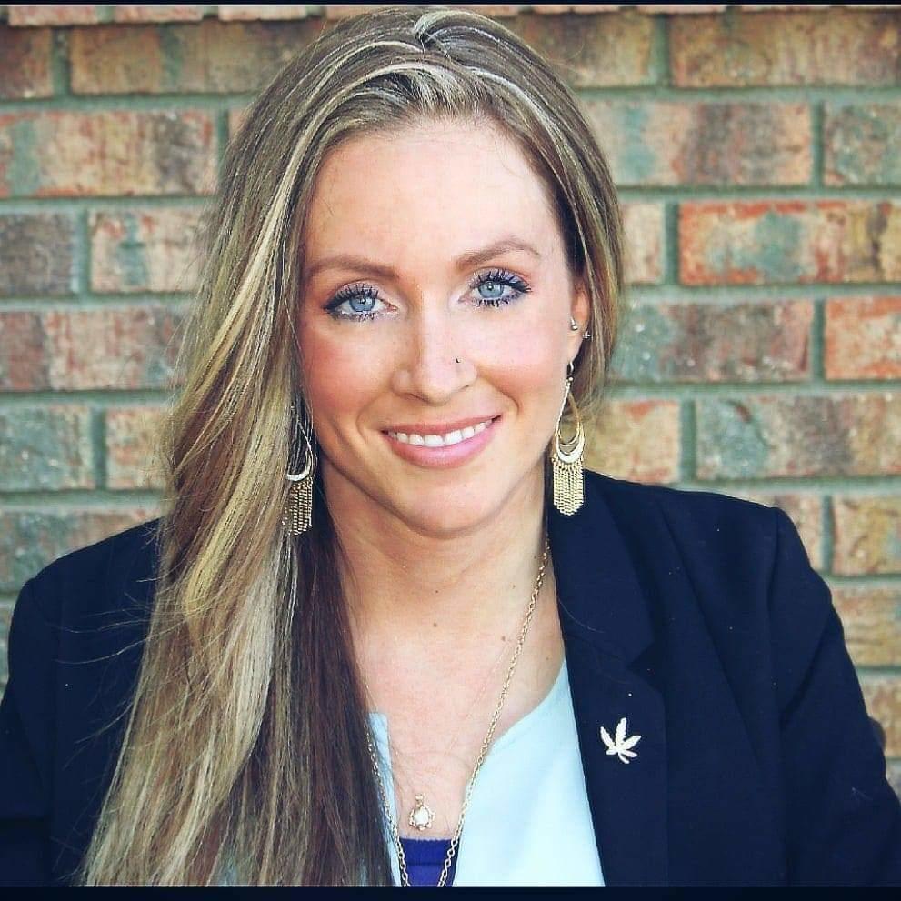 A fair skinned woman with blond long, straight hair and blue eyes, smiling, wearing a black blazer and blue undershirt