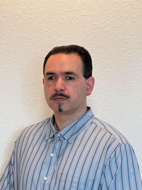 A medium skin toned man with black hair, brown eyes, a mustache with a goatee, wearing a button up stripped long sleeved shirt.