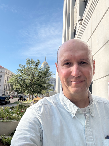 Bald man in a white button down shirt standing on the streets of Denver smiling.