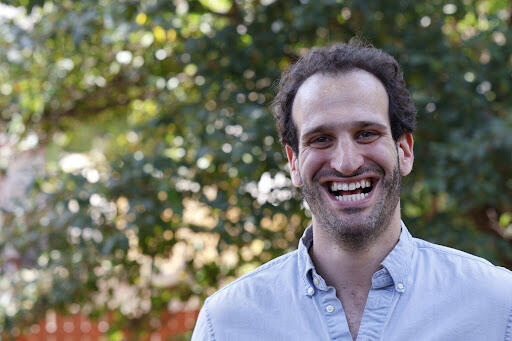 Man with dark brown hair wearing a light blue button down shirt standing in front of green leaf trees smiling