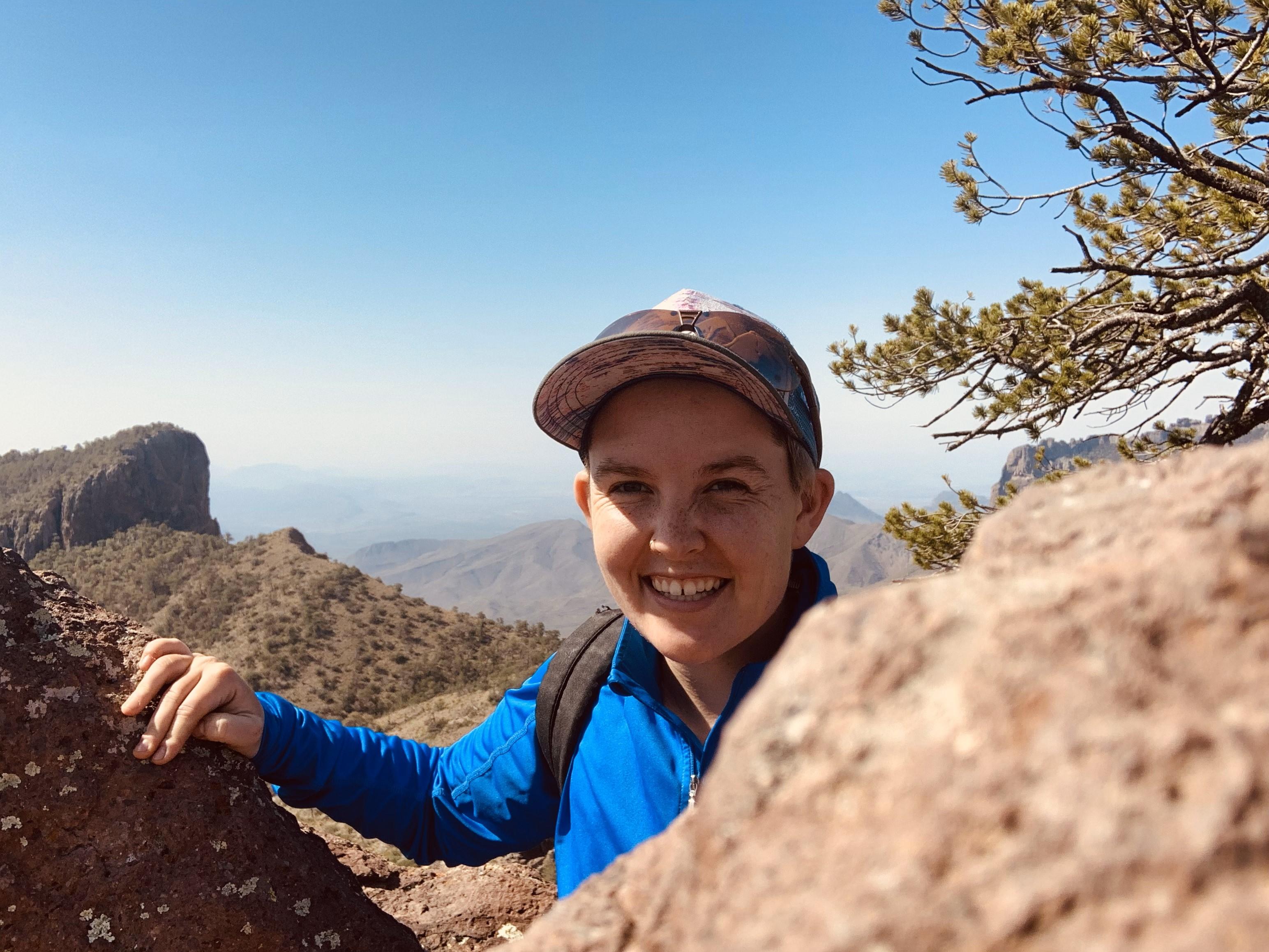 A smiling light skinned person with a hat on, blue long sleeve shirt, a backpack, who is rock climbing