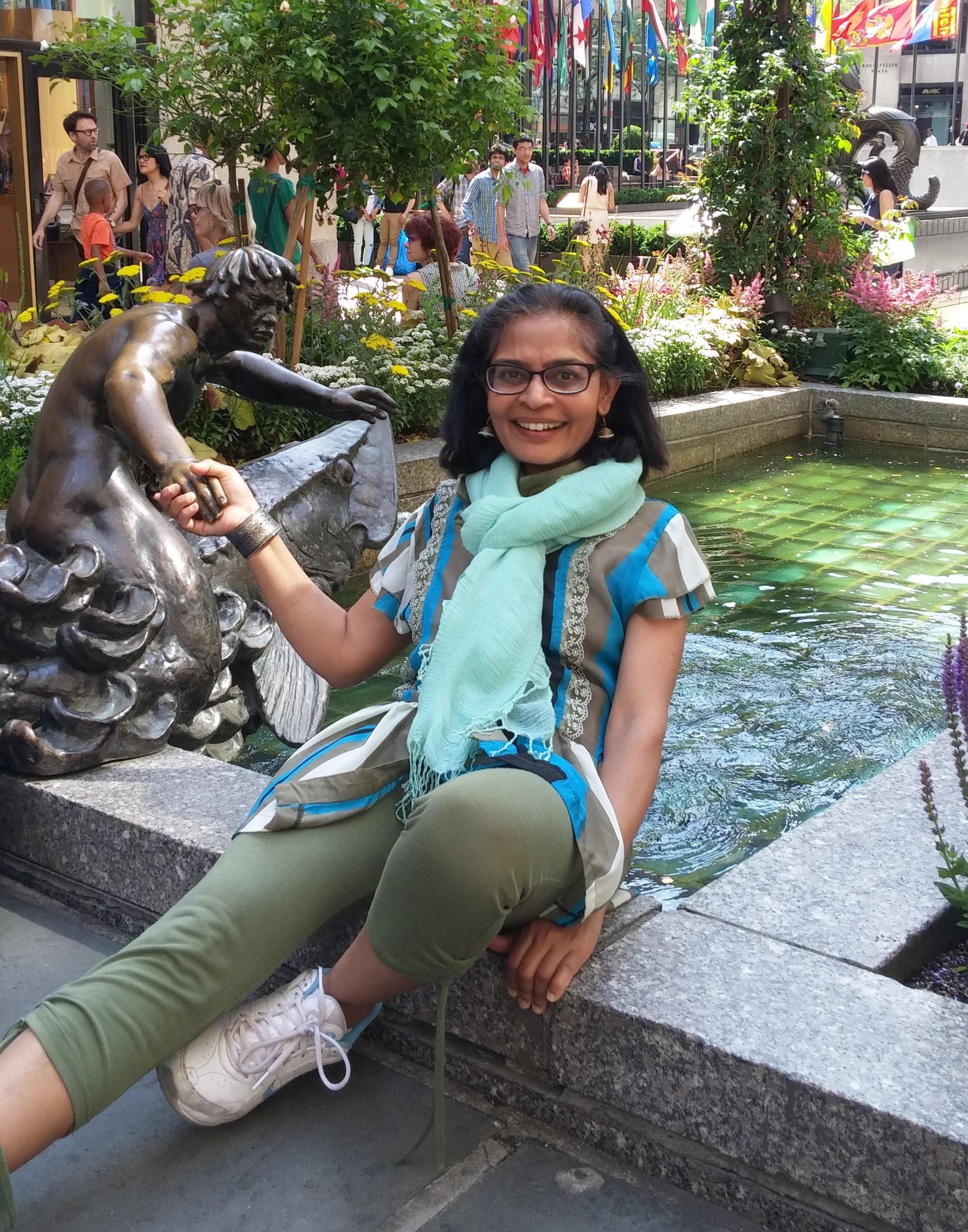 A dark complexed Indian woman with black hair shoulder length, brown eyes, smiling, wearing green pants, a light blue scarf, and a multi-patterned shirt, holding hands with a statue.