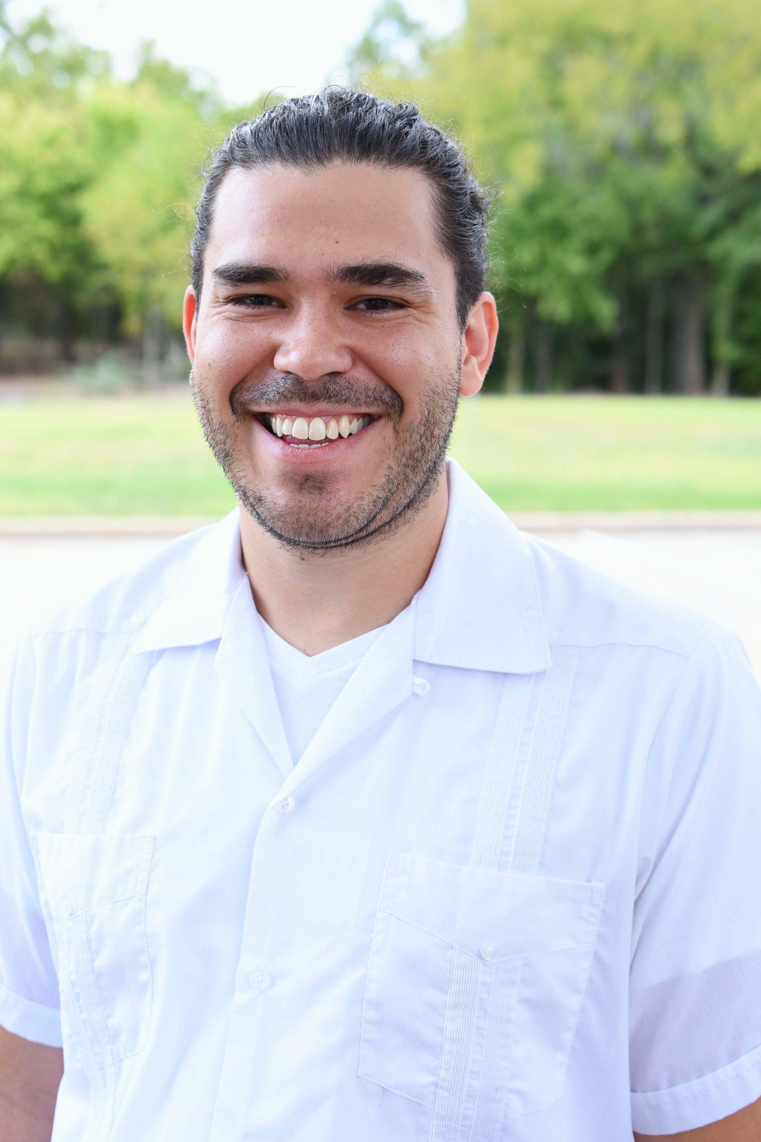 A profile photo of Mateo Clarke. Mateo is a person with dark brown hair pulled back behind his head. He is wearing a white collared, short-sleeve shirt and is standing in front of green trees and grass.
