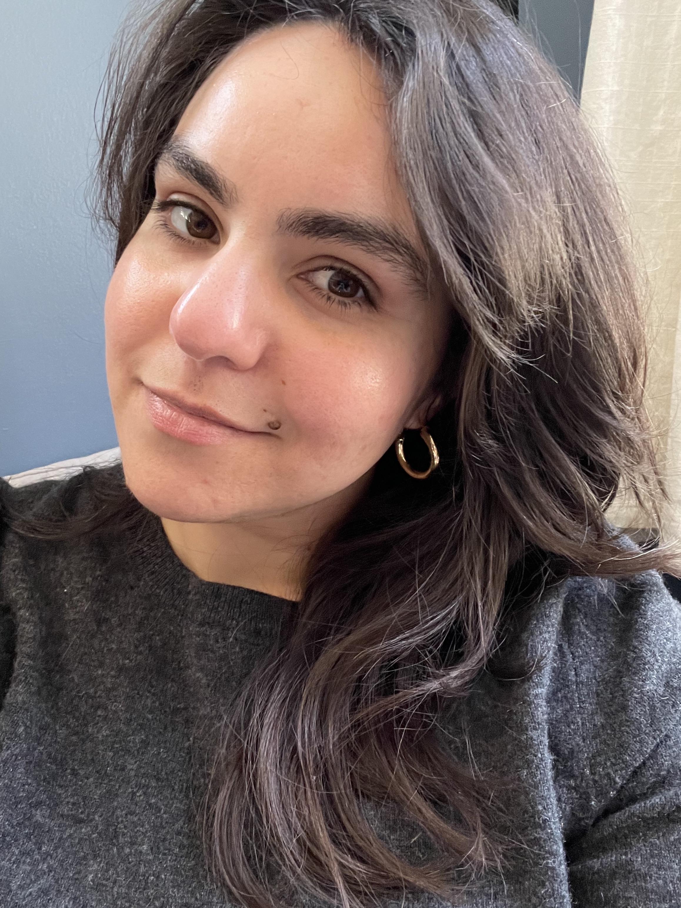 A profile image of Mina Farzad. Mina has long dark hair and a grey sweater. She is tilting her head to one side and is standing in front of a blue wall.