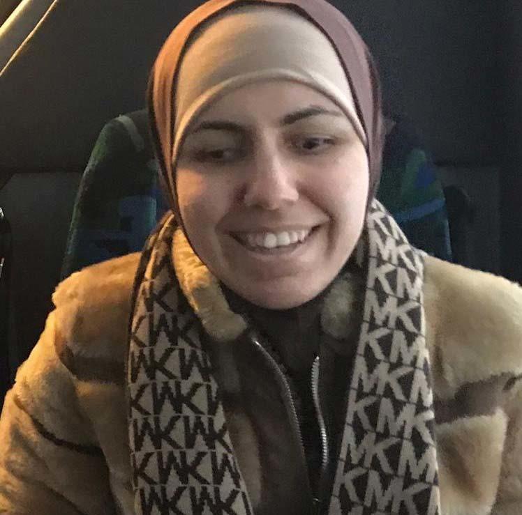 An Iraqi woman with a medium complextion, brown eyes, smiling, wearing a brown coat, with a scarf that is brown and tan with the letters M and K