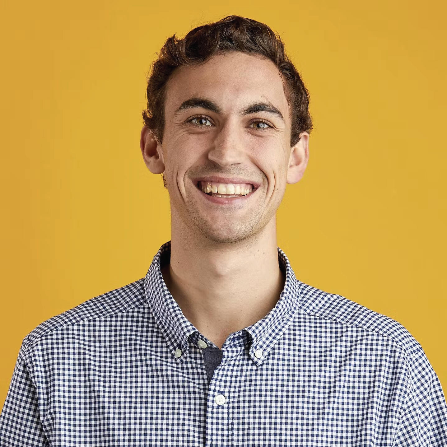 Tristan is a person with short brown hair and brown eyes. They are smiling and wearing a blue checkered, button-up shirt. They are standing in front of a yellow background.