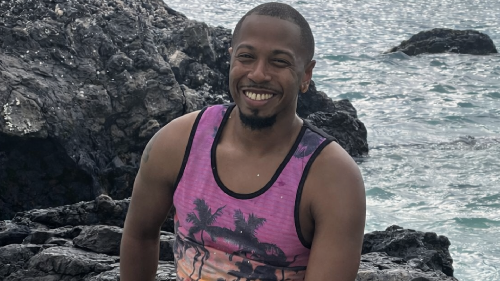 A smiling man sits on dark volcanic rocks by the ocean as a wave splashes up in front of him.