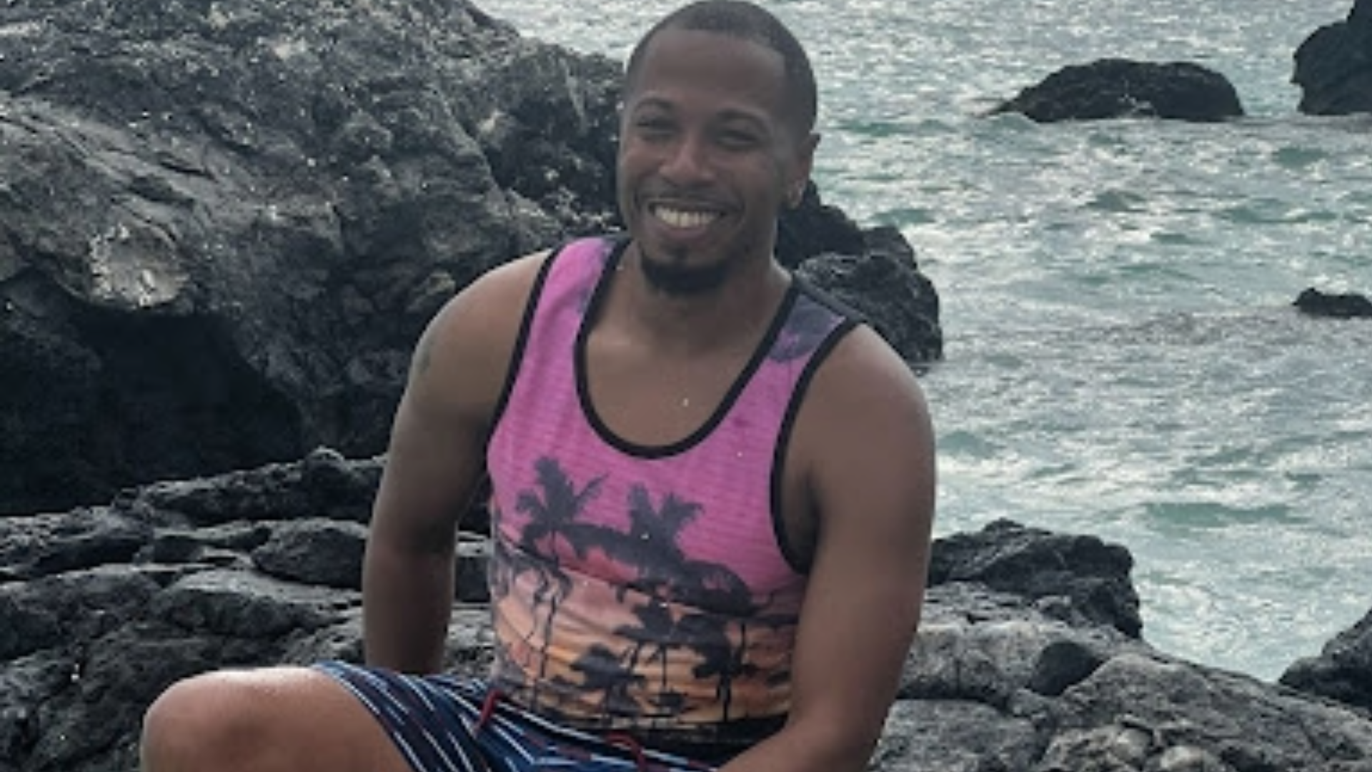 A smiling man sits on dark volcanic rocks by the ocean as a wave splashes up in front of him.