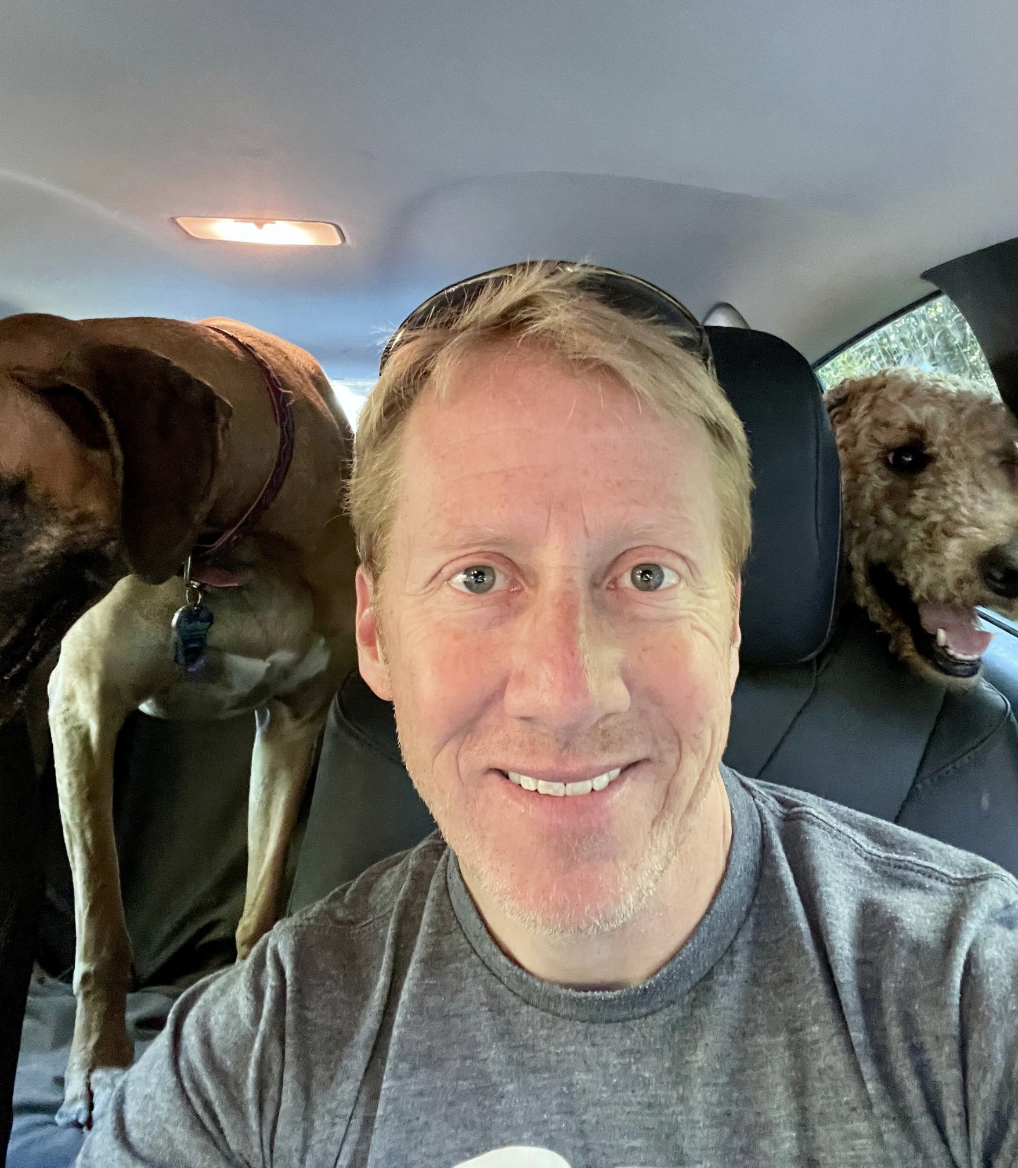 A white man with light skin, blue eyes, and blonde hair looks into the camera and smiles warmly. He is sitting in the front seat of a car, his dogs smile happily in the back seat.