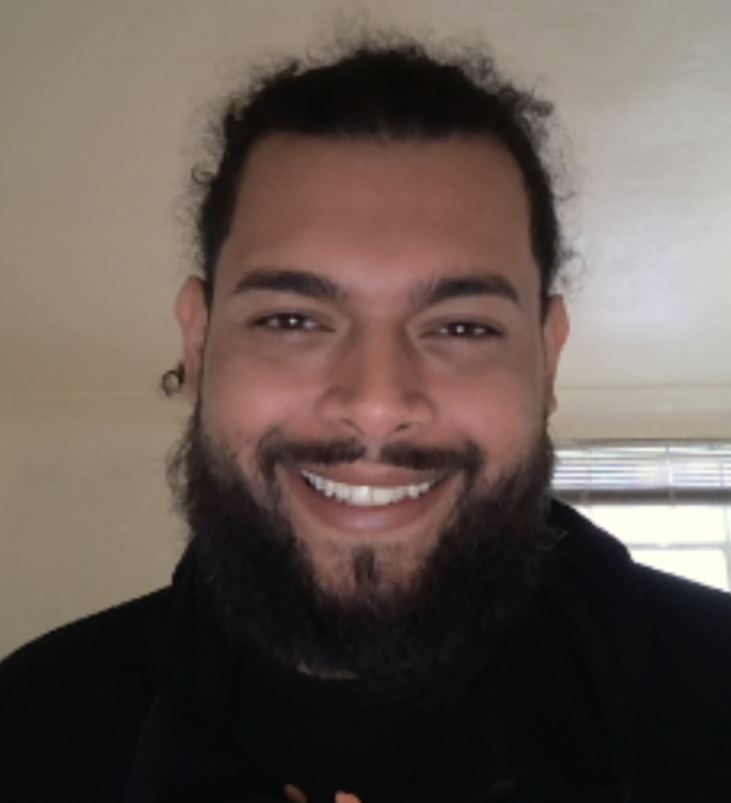 A man with light brown skin and black curly hair pulled back and a thick beard looks directly into the camera and smiles happily. 