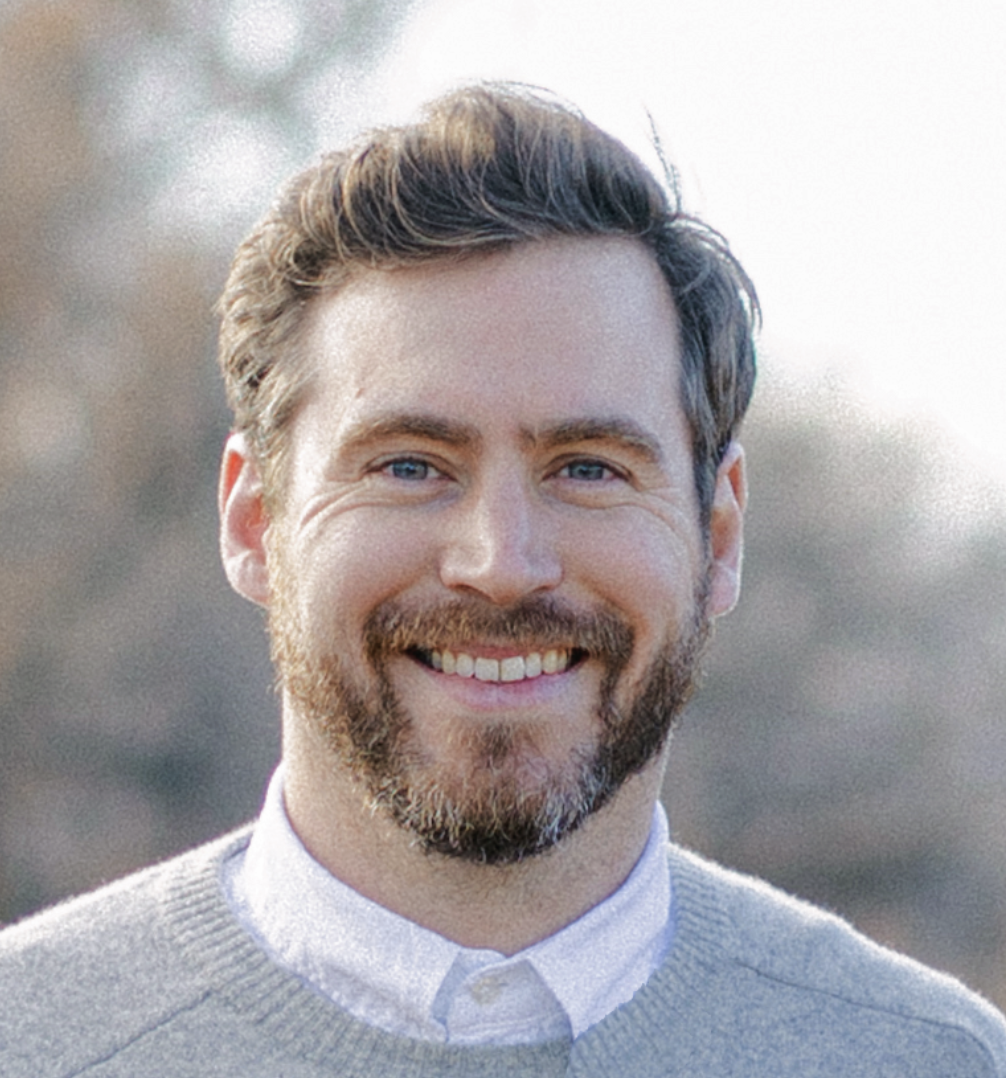 A white man with blue eyes and short brown hair with facial hair peppered with a few gray hairs looks directly into the camera and smiles. He is wearing a white collared shirt under a gray sweater.