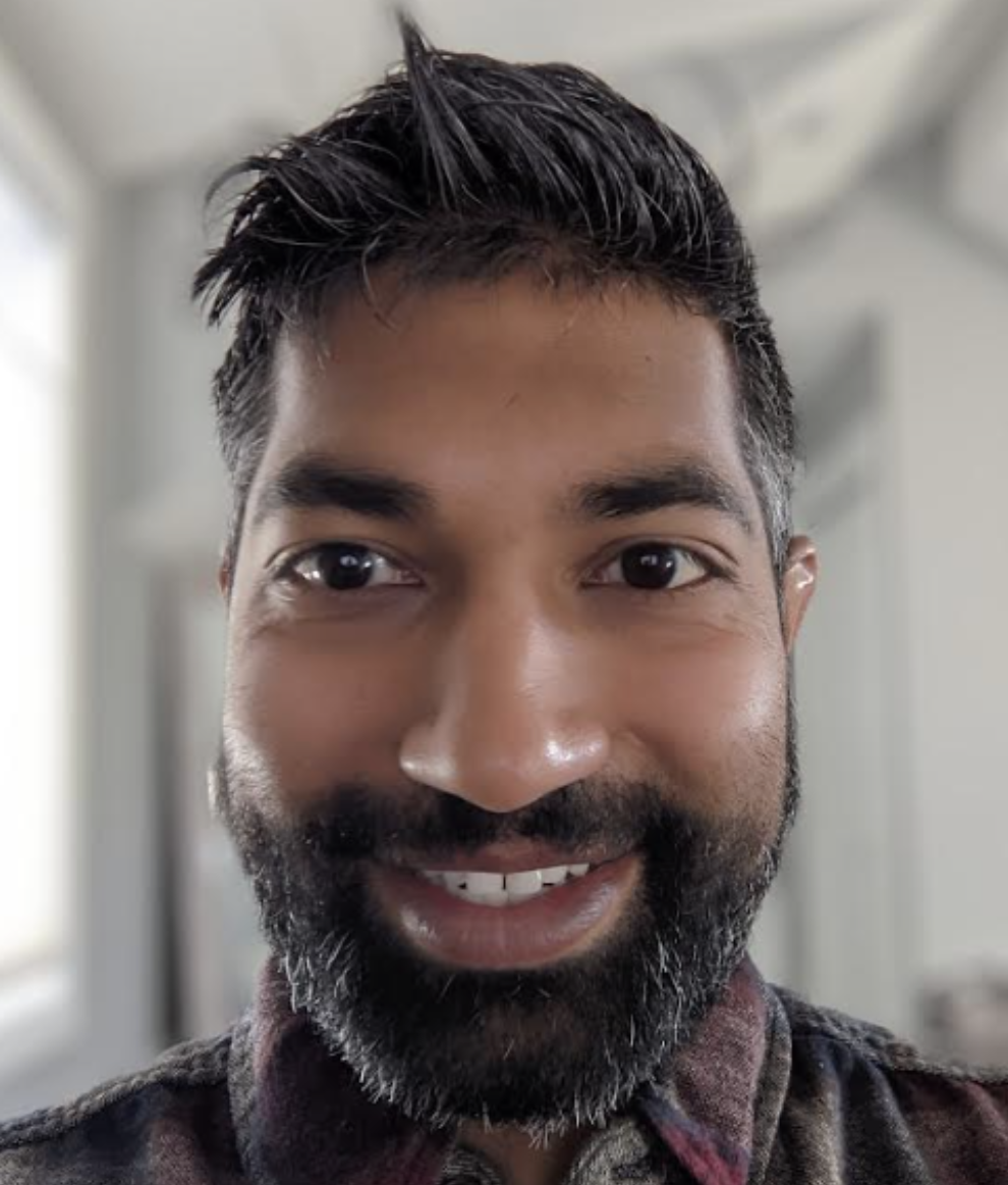 A man with brown skin, dark brown eyes, and short black hair with facial hair peppered with a few gray hairs looks directly into the camera and smiles warmly.