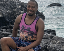 A smiling man sits on dark volcanic rocks by the ocean as a wave splashes up in front of him.
