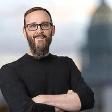 professional portrait of a man with a beard and glasses, posing with his arms crossed against a blurred architectural background.
