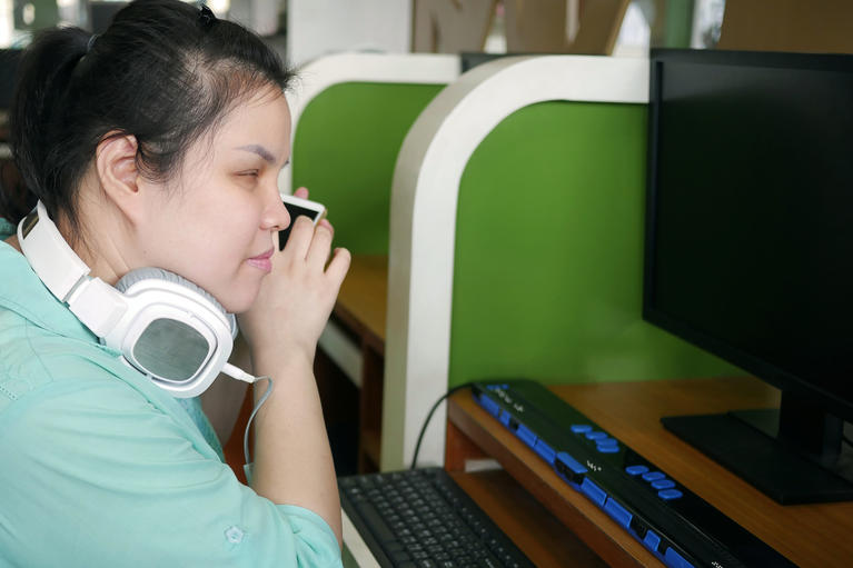 Asian young blind person woman with headphone using smart phone with voice assistive technology for disabilities persons in workplace with computer and braille display