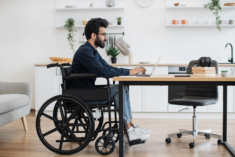 Man in wheelchair using laptop at desk in bright kitchen